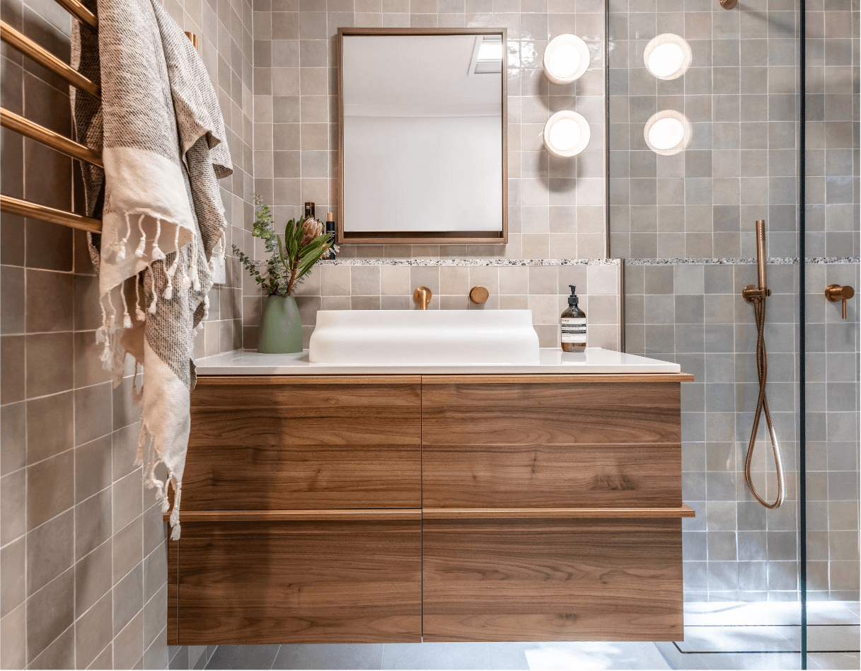 A multi-color gray tiled bathroom with large wood vanity and a walk-in tiled shower to the right with bronze-colored fixtures.