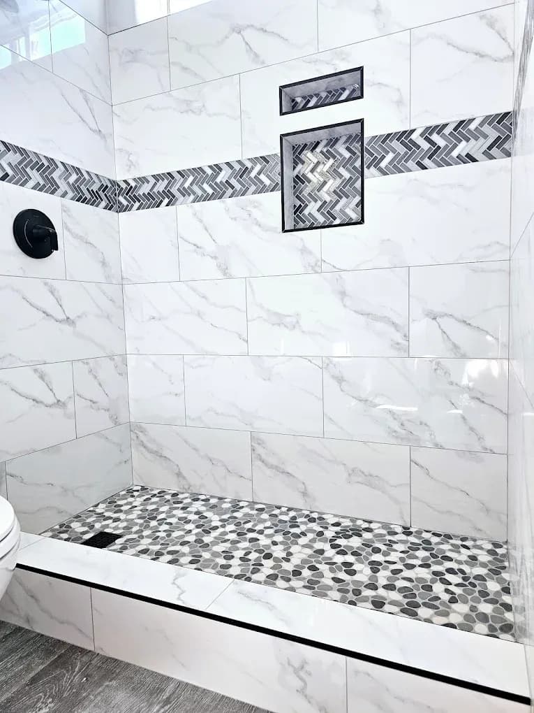 A step-in shower with white and gray marbled tile walls and black, white, and gray stone mosaic accents.