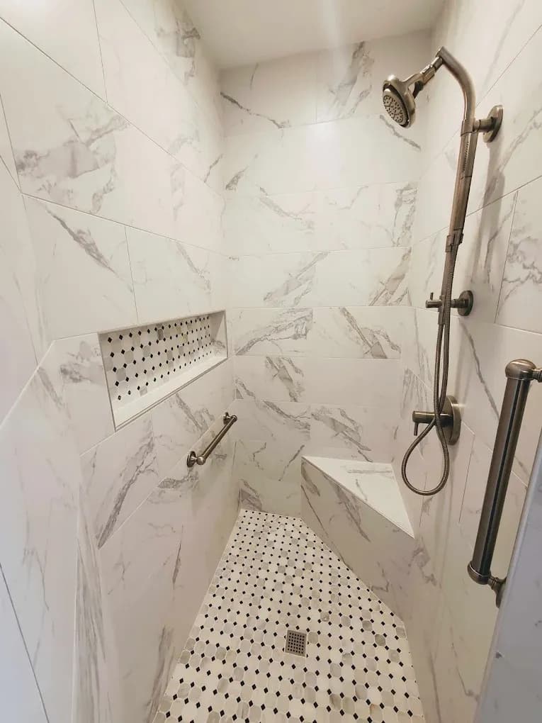A shower with white and gray marbled stone walls, black and white tile floor and wall caddy, built-in corner seat, and vertical grab bar.