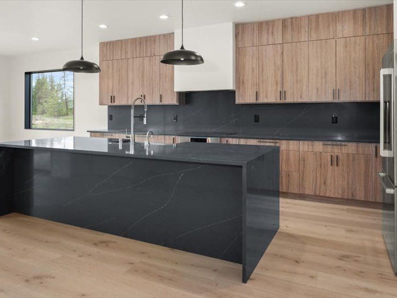 A kitchen with black marbled countertops and waterfall-style island, medium brown cabinets, and LUX vinyl plank flooring.