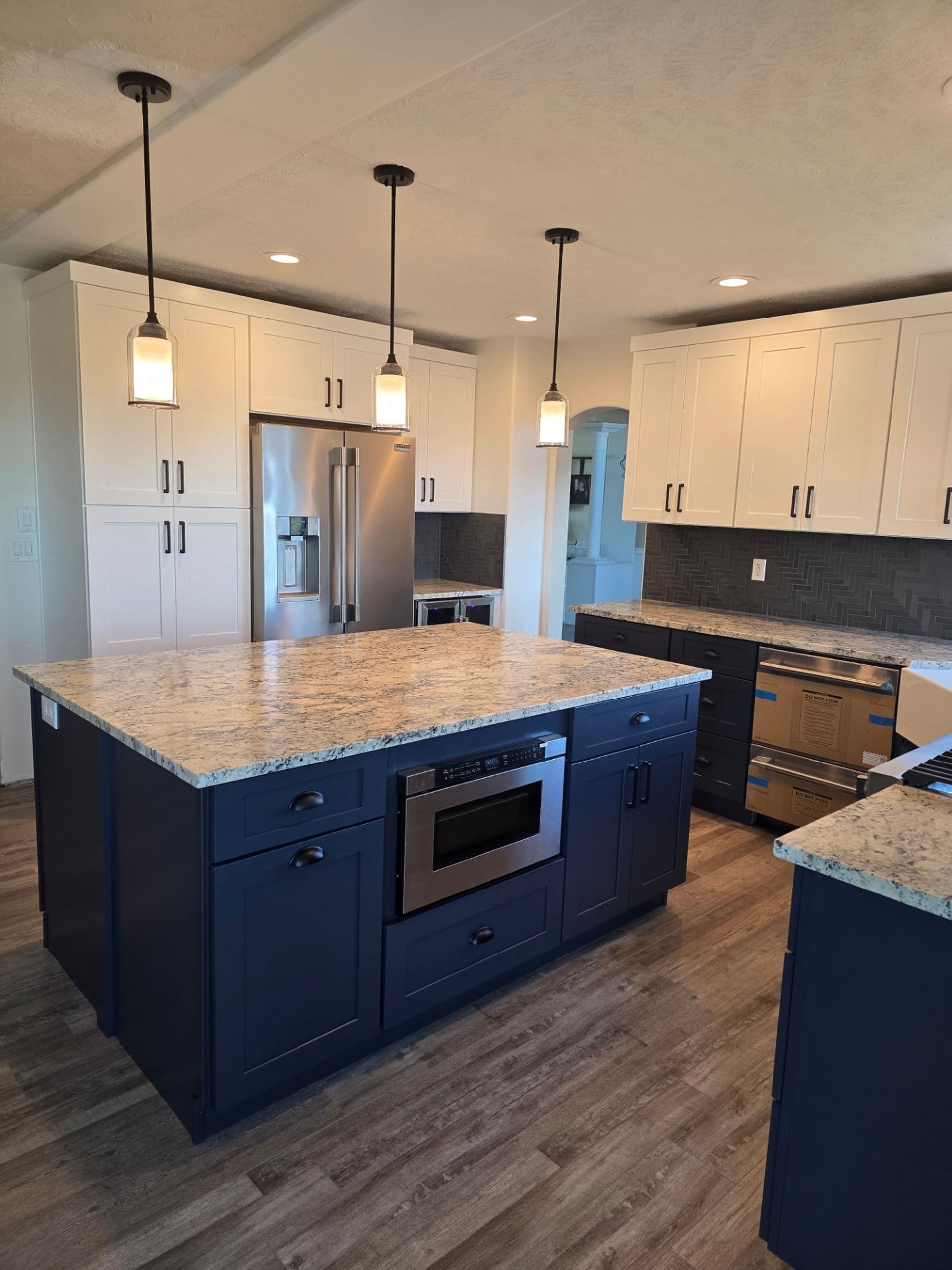 A kitchen with white cabinets, large central island with dark blue cabinets and built-in microwave, and gray marbled countertops.