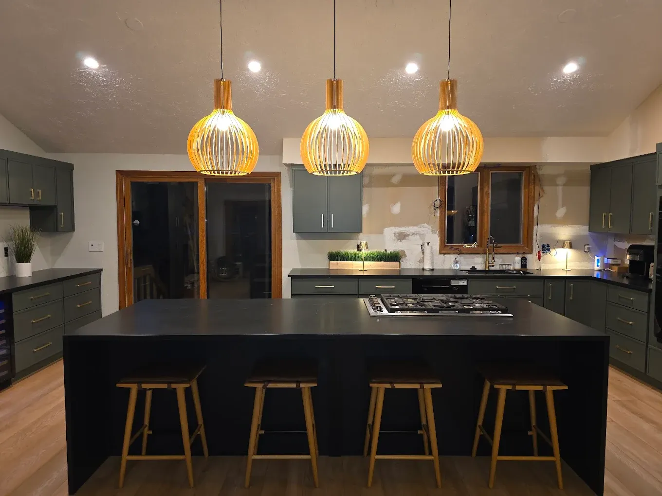 A kitchen with dark gray-green cabinets and a large central island with black stone waterfall countertop and bar stool seating.